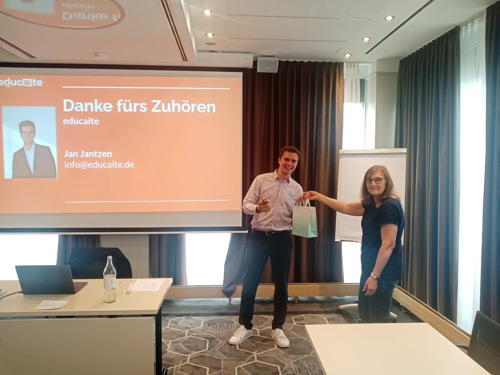 Monika Holder überreicht Referent Jan Jantzen eine Geschenktüte, dahinter die Abschlussfolie „Danke fürs Zuhören“ mit educaite-Logo