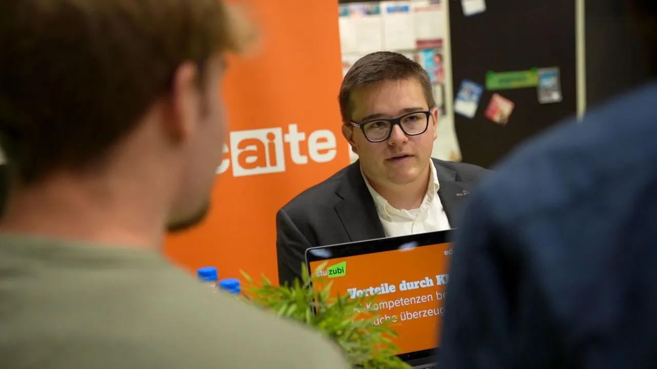 Beratungsgespräch am educaite-Stand: Ein Berater im Sakko spricht mit zwei Messebesuchern, daneben das orange educaite-Banner und ein Laptop mit der Folie „Vorteile durch KI“